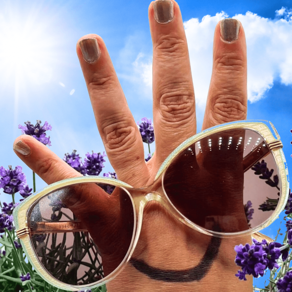 Das Foto zeigt im Hintergrund ein Lavendelfeld vor strahlend-blauem Himmel. Das Hauptmotiv ist eine Hand mit gespreizten Fingern, die eine Sonnenbrille trägt und auf die unten ein lächelnder Mund gemalt ist. Es ist ein sehr sommerliches und fröhliches Motiv, das die Lust am Reisen verdeutlichen soll.